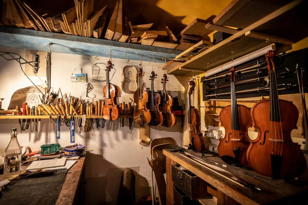 Laboratorio liutaio: violini, viole, violoncelli e archi appesi. Legname e attrezzi da lavoro, con un vasetto di kerria lacca.