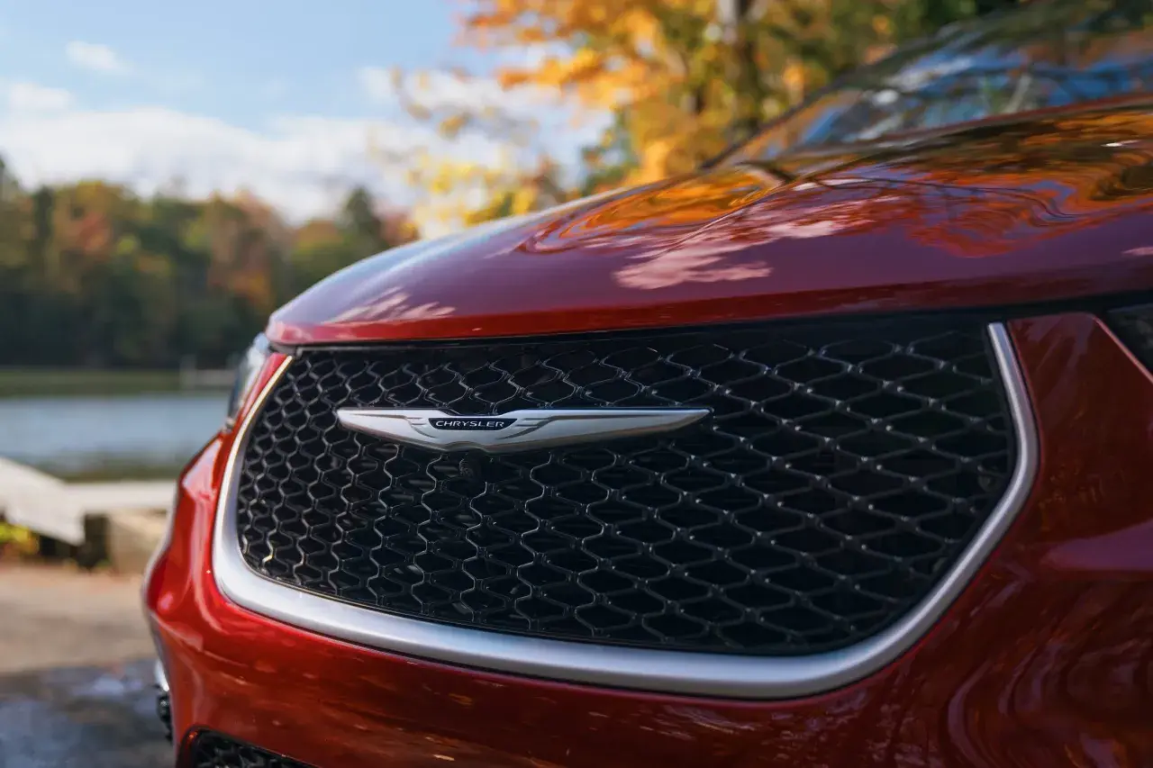 En röd Chrysler-bil med sitt ikoniska märke framtill, parkerad vid en sjö under höstens färger.