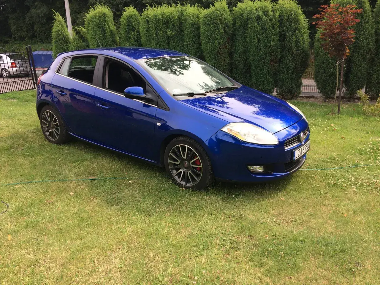Błękitny Fiat Bravo 1.4 T-Jet zaparkowany na trawniku, za nim żywopłot.