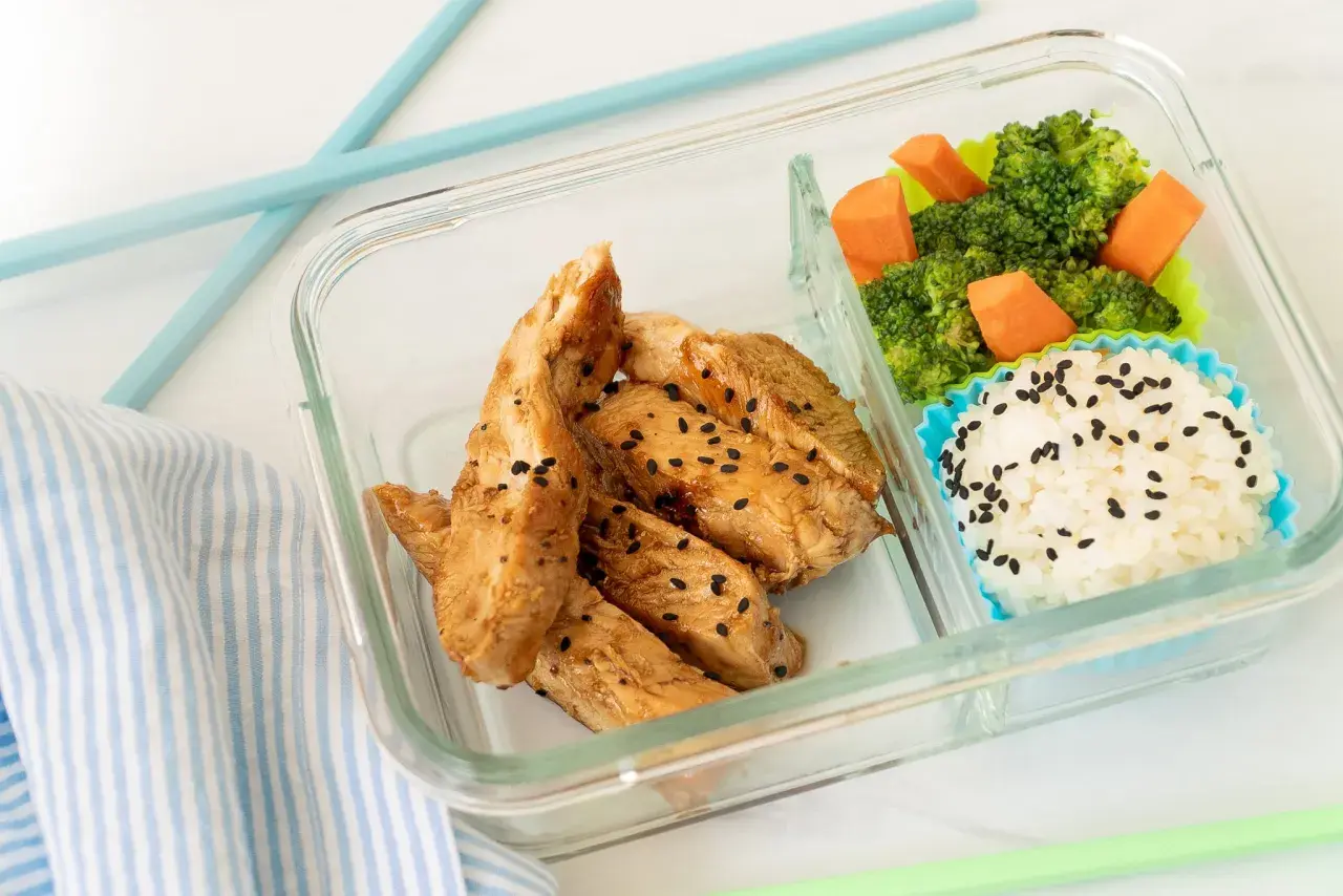 A delicious chicken bento box with teriyaki chicken strips, white rice sprinkled with black sesame seeds, and steamed broccoli with carrots.