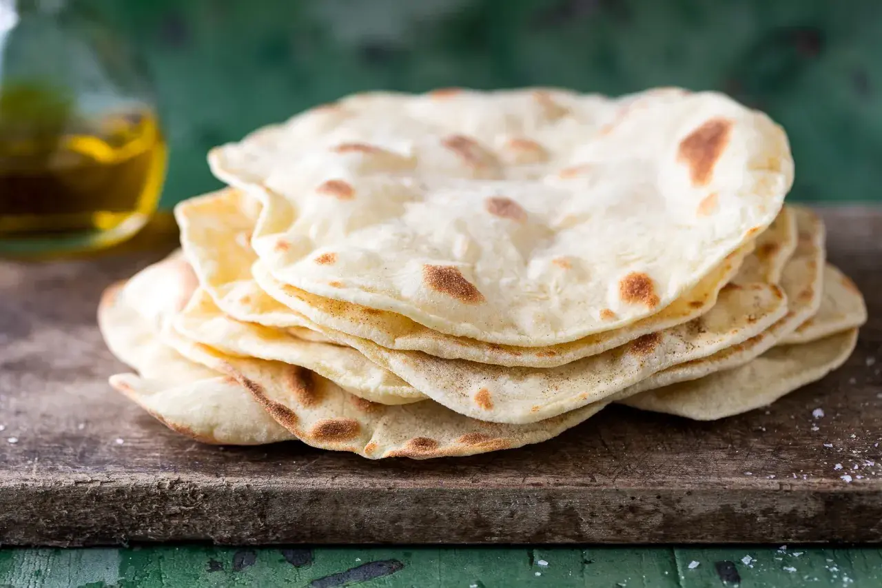 Pila di pane senza lievito, cotto su una tavola di legno grezzo, con un tocco di sale e una bottiglia d'olio sullo sfondo.
