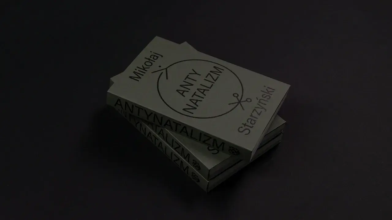 Stos książek z napisem "ANTY NATALIZM" i symbolem nożyczek. Dzieło Mikołaja Starzyńskiego.