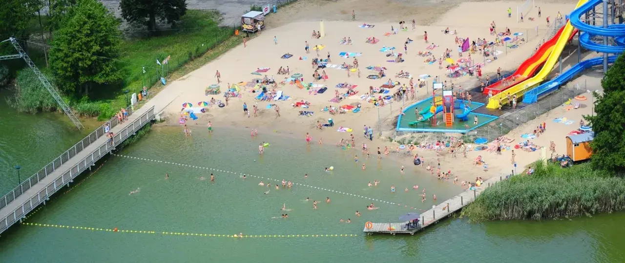 Wakacyjny dzień nad jeziorem margonińskim. Ludzie opalają się na plaży, kąpią w wodzie i korzystają ze zjeżdżalni wodnych.