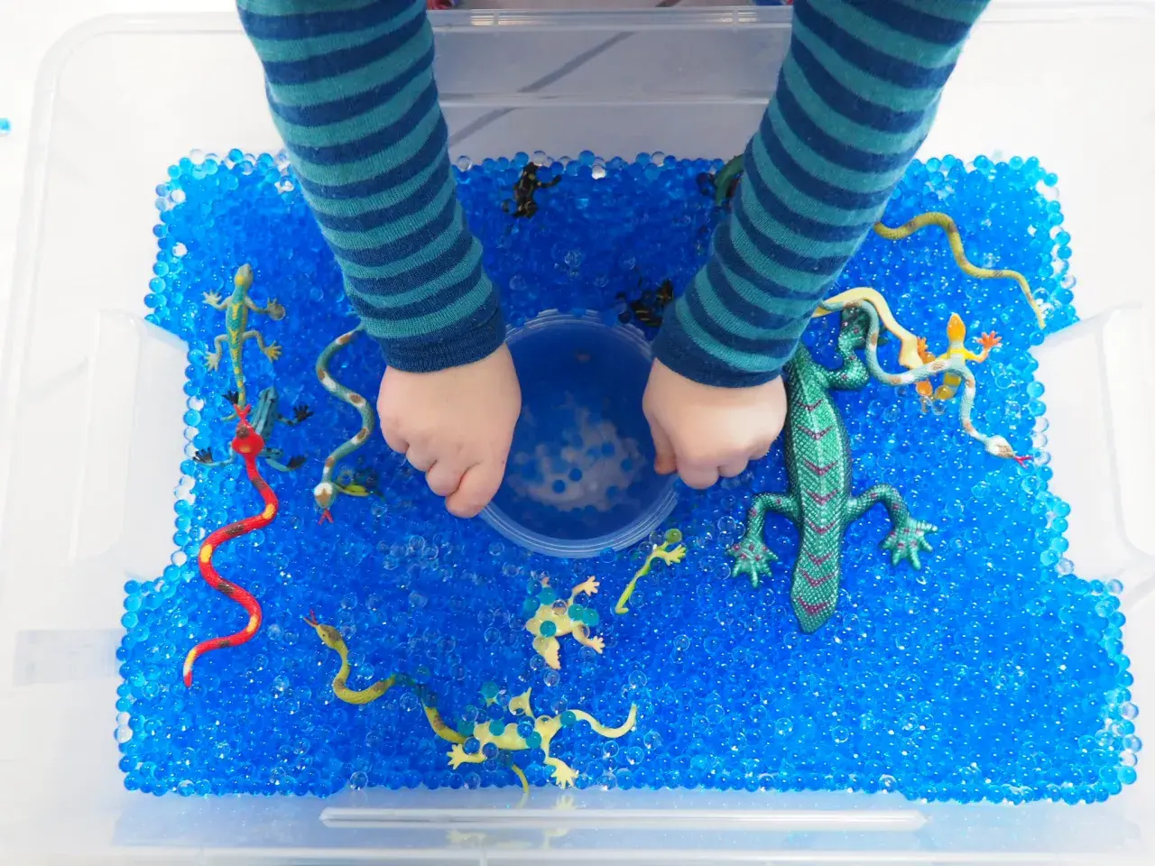 Kinderhände erkunden eine Kiste voller blauer Wasserperlen mit Spielzeug-Echsen und Schlangen. Tolle Wasserperlen Ideen für sensorisches Spiel!