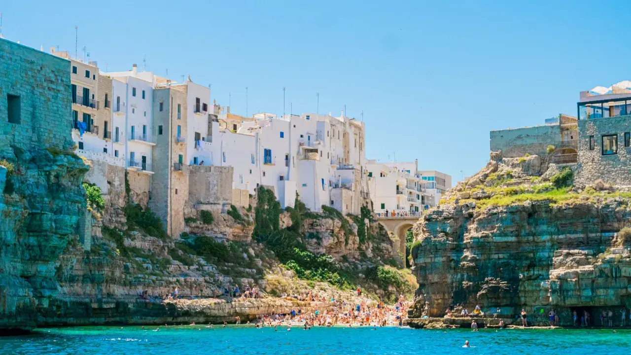 La famosa spiaggia di Polignano a Mare, con case bianche aggrappate alle scogliere e bagnanti che si godono il mare cristallino.