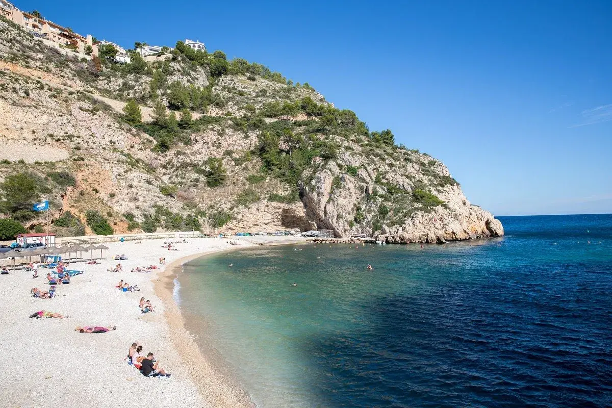 Gente disfrutando del sol en una playa de guijarros en la **Costa Blanca**, con aguas turquesas y un acantilado rocoso.