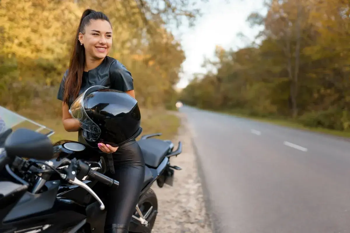 Uśmiechnięta kobieta w skórzanym stroju trzyma kask obok czarnego motocykla. Idealne motory dla kobiet na jesienną przejażdżkę.