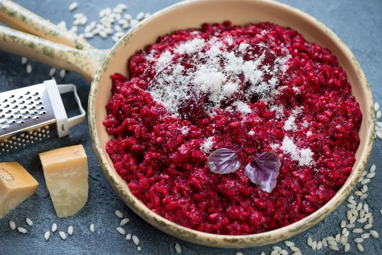 Risotto rosso vibrante, guarnito con parmigiano grattugiato e basilico. Accanto, ingredienti per un perfetto vino rosso per risotto.