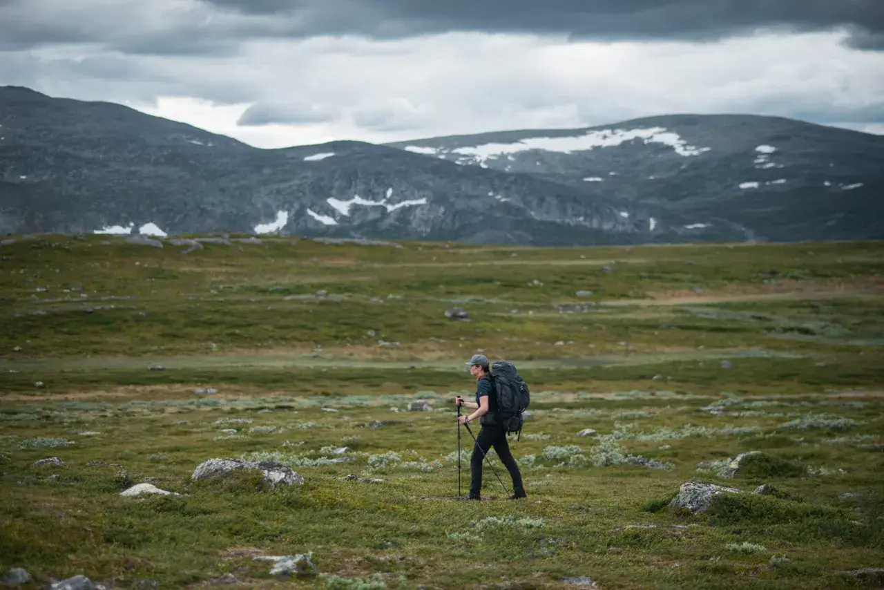 Vandrare med ryggsäck och stavar går över kalfjäll med snöfläckar på berg i bakgrunden. En helags karta skulle visa vägen.