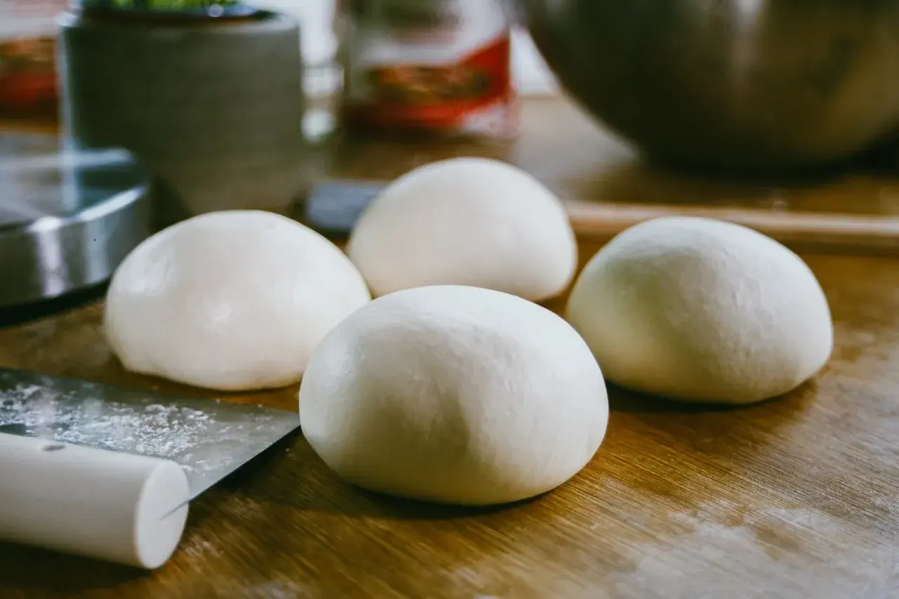 Cztery kulki surowego ciasta na pizzę leżą na drewnianym blacie obok noża.