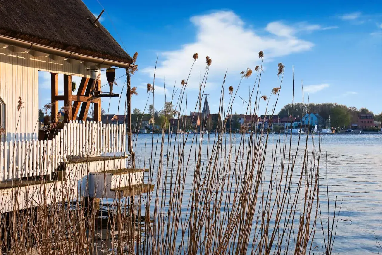 Reetgedecktes Haus am Plauer See mit Blick auf die Stadt und den Kirchturm. Schilf wiegt sich im Wasser.