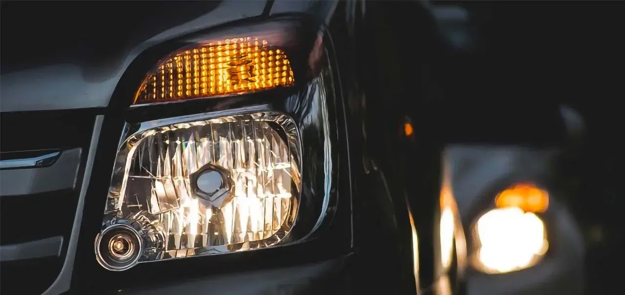 Luces cruce de un coche, con el intermitente naranja encendido y el faro principal iluminando. Otro coche se ve al fondo.