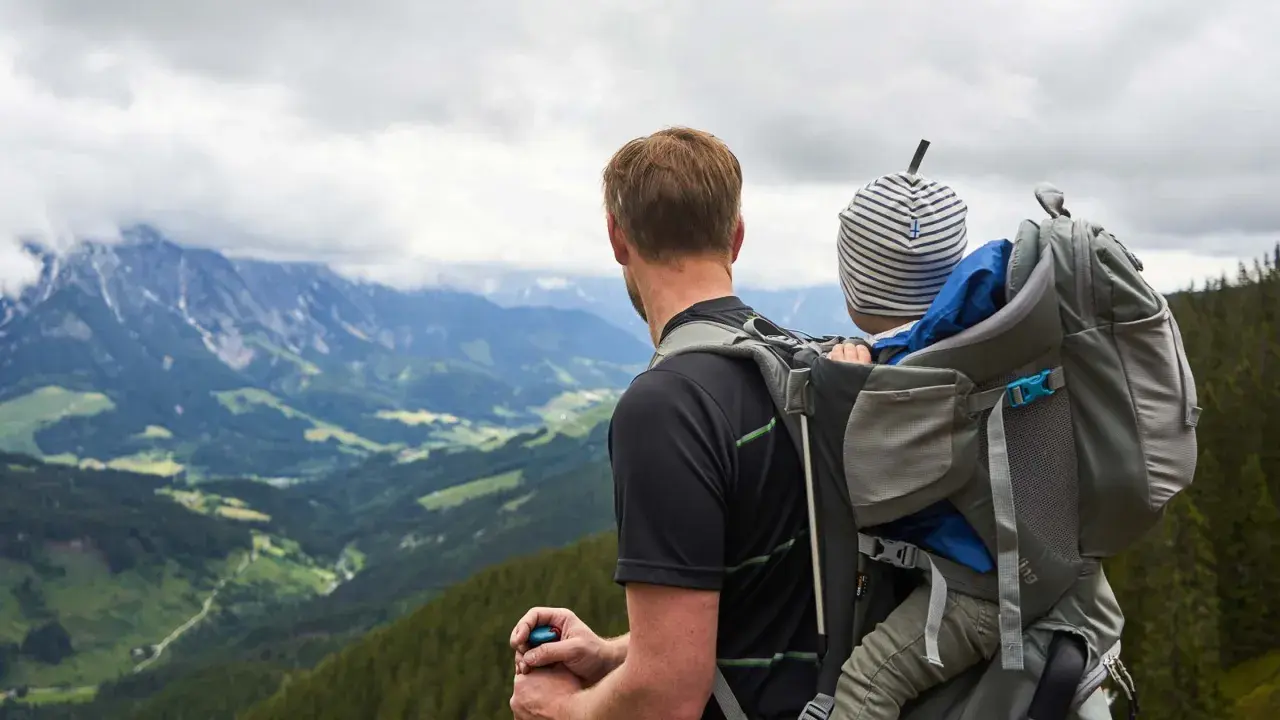 Mann mit Kind auf dem Rücken in einer Kraxe, blickt auf bergige Landschaft.