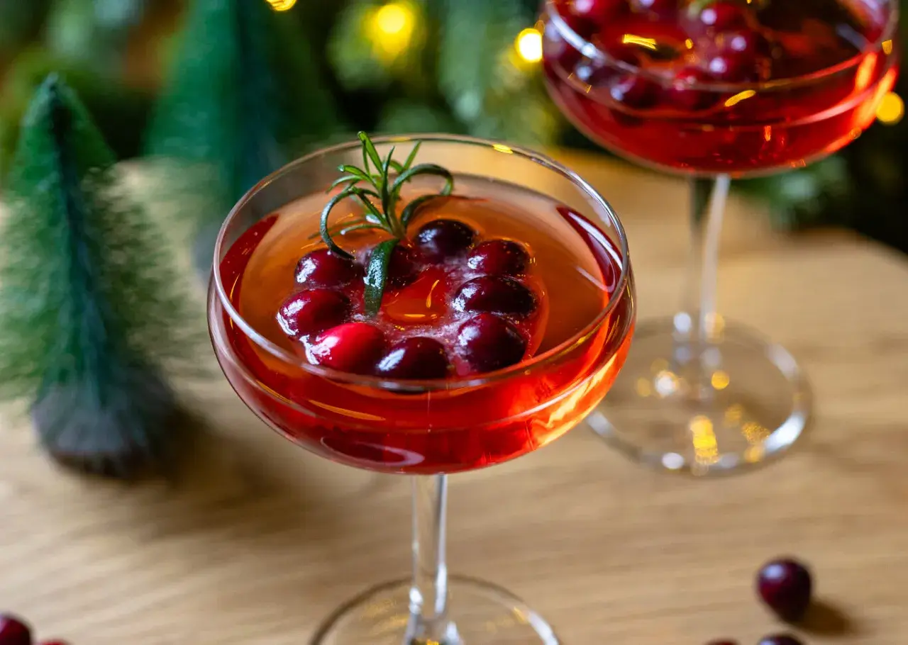 Festliche Aperol-Cocktails mit Cranberries und Rosmarin, perfekt für Aperol Weihnachten. Kleine Bäume und Lichter im Hintergrund.