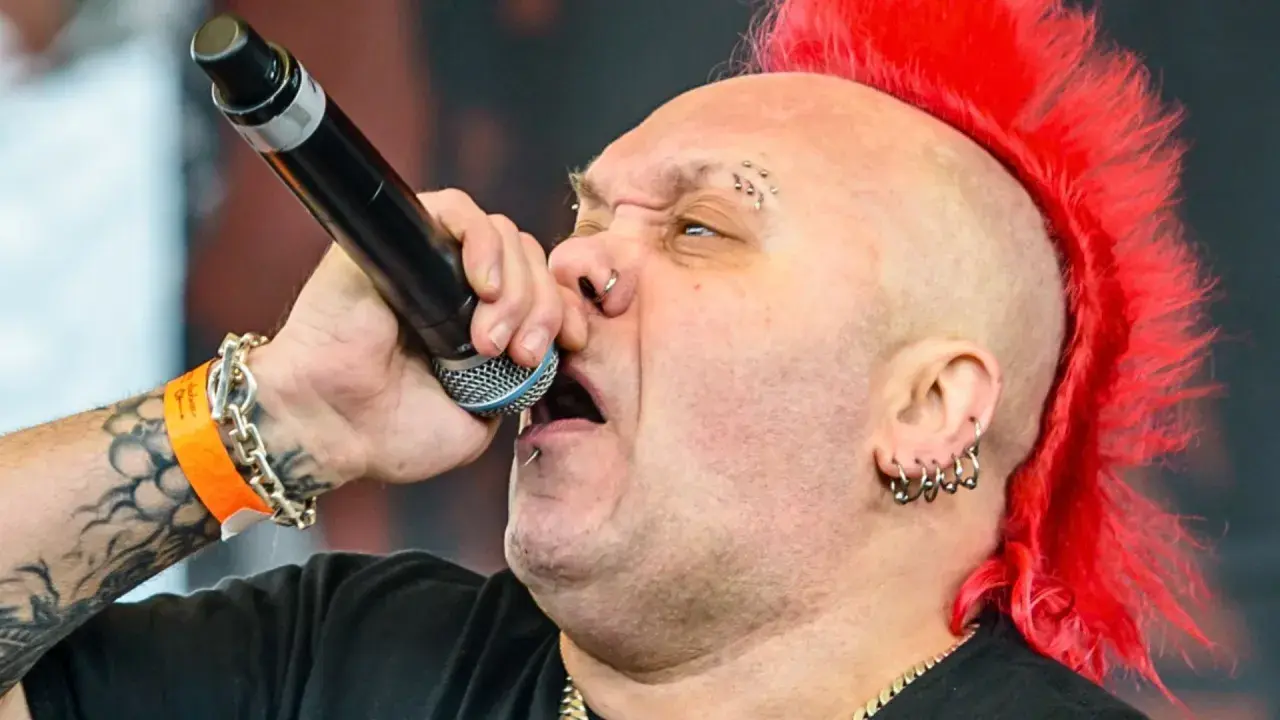 Wokalista z irokezem śpiewa do mikrofonu. Widać tatuaże, kolczyki i opaskę festiwalową. Jego styl sugeruje, że nie dał się łatwo wykorzystać.