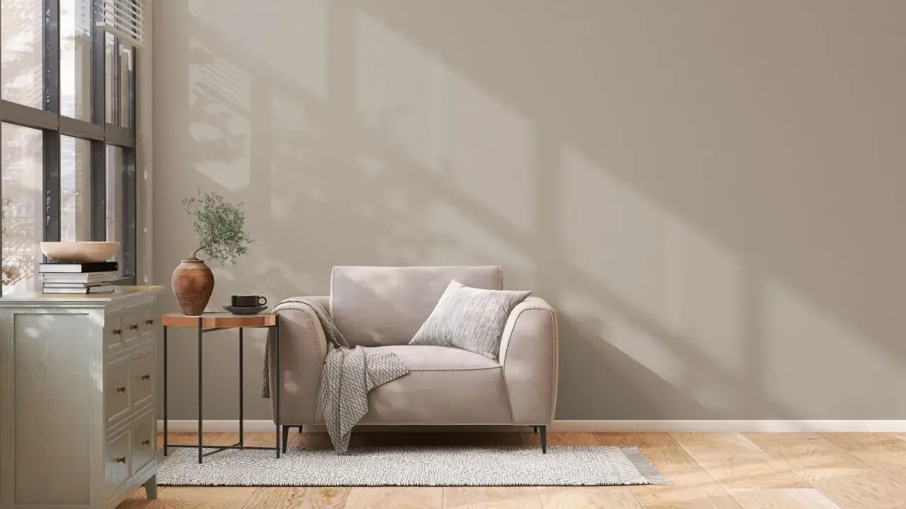 Un sillón beige con manta y cojín, junto a una mesita auxiliar con jarrón y taza. La pared tiene unos colores de pintura neutros, con sombras de luz.