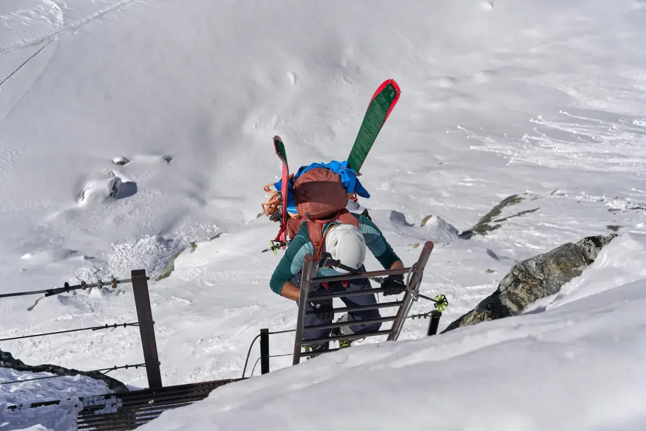 Escalade d'une échelle sur un flanc de montagne enneigé, skis sur le dos. L'ascension vers un sommet 4000 des Alpes, une aventure pas si facile.