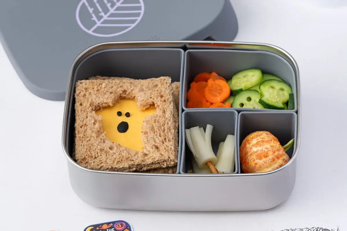 A spooky Halloween bento box with a ghost-shaped cheese sandwich, carrot slices, cucumber rounds, and a peeled tangerine.