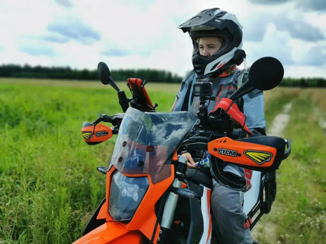 Motocyklista w kasku siedzi na pomarańczowym motocyklu w terenie, na tle zielonego pola i nieba.