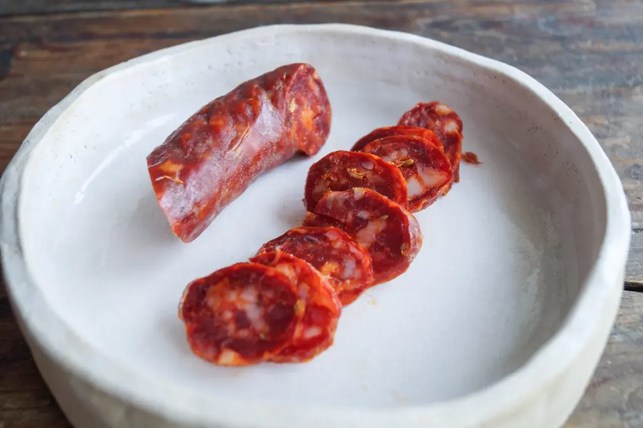 Ein Stück und Scheiben einer würzigen Salsiccia Wurst liegen in einer weißen Schale auf einem Holztisch.