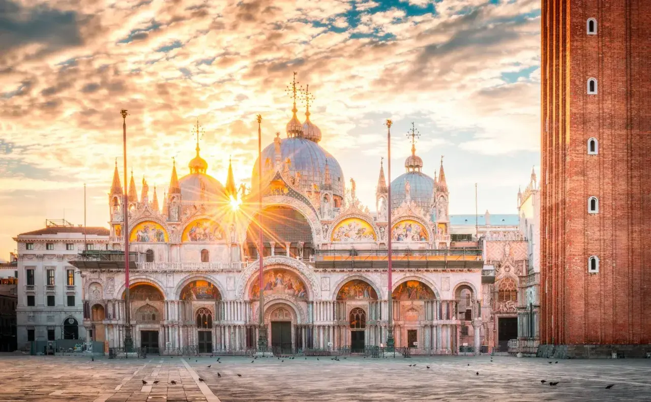 Wschód słońca nad Bazyliką św. Marka na Piazza San Marco. Złote promienie przebijają się przez chmury, oświetlając wenecką architekturę.