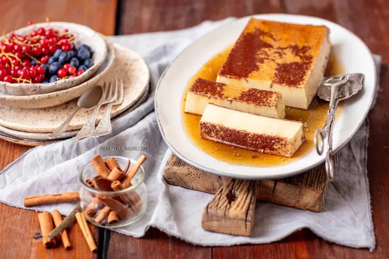 Pyszny creme caramel na talerzu, polany syropem. Obok miseczka z owocami i laska cynamonu.