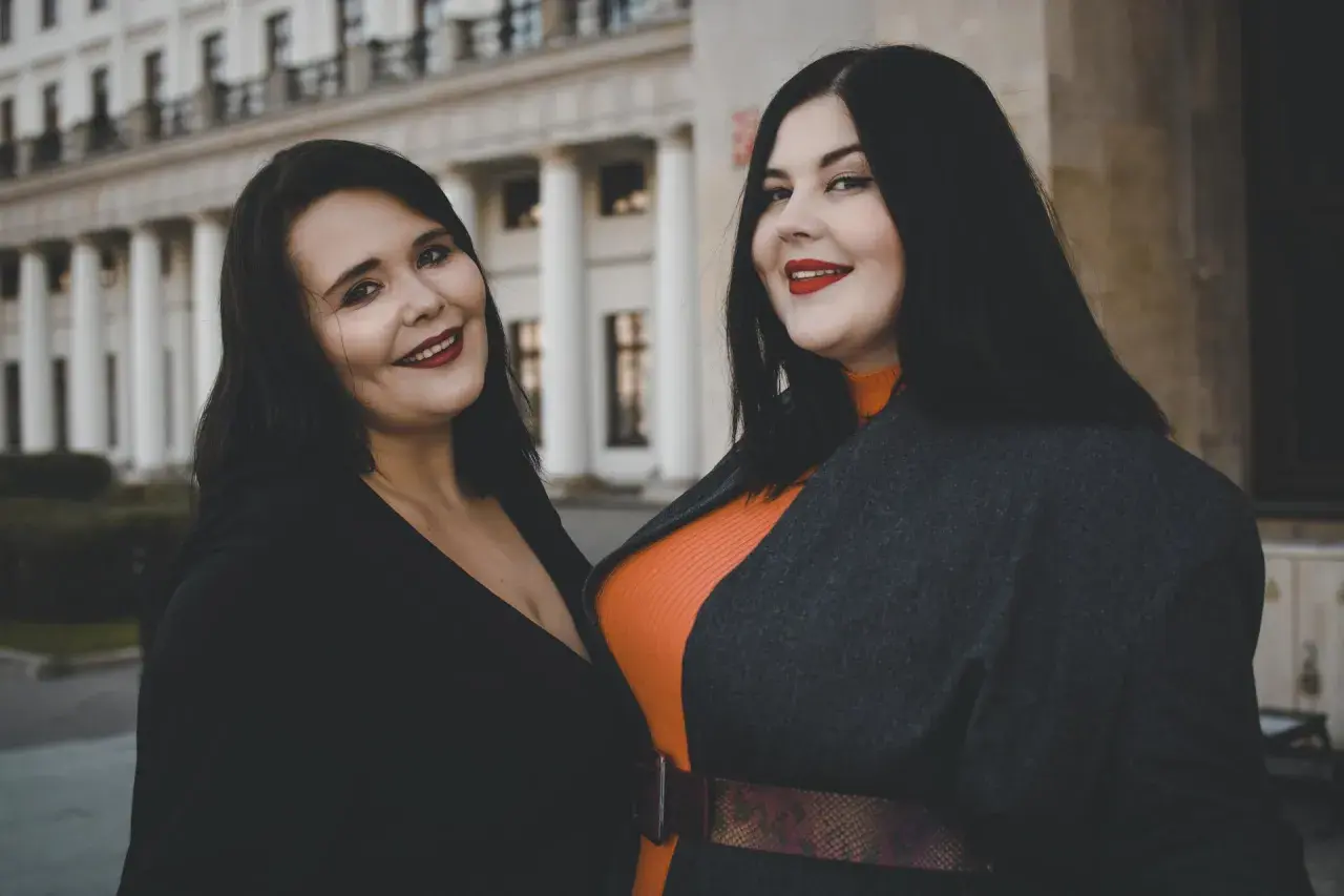 Dwie uśmiechnięte kobiety, uosobienie body positivity, pozują przed budynkiem z kolumnami.