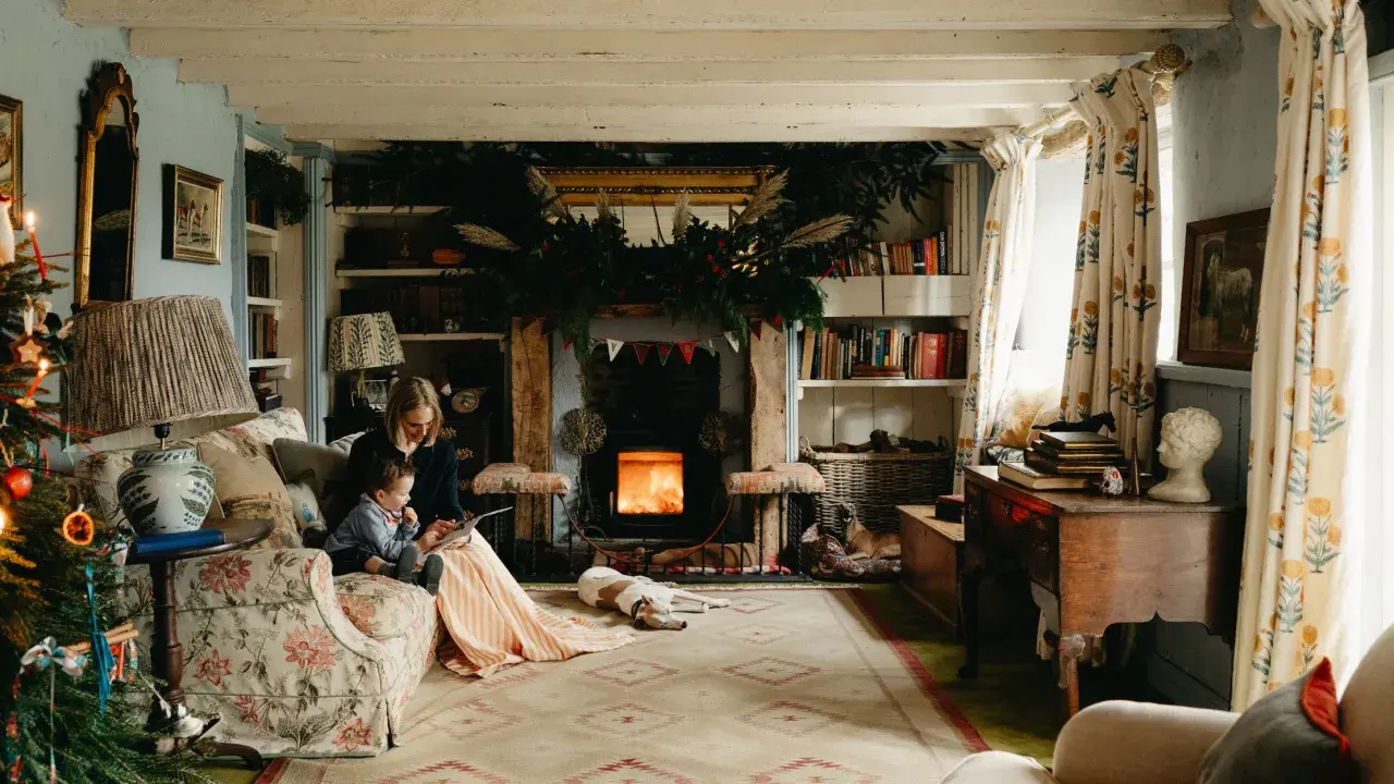 Cozy cottagecore house scene with a woman and child reading by a fireplace, dogs resting, and festive Christmas decorations.