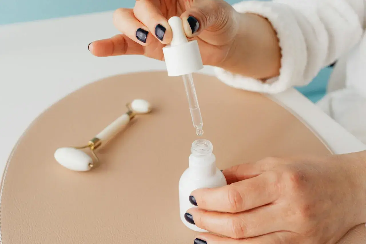 Dłoń z ciemnym lakierem do paznokci trzyma pipetkę z serum. Obok leży jadeitowy roller. Pielęgnacja: niacynamid z czym nie łączyć.