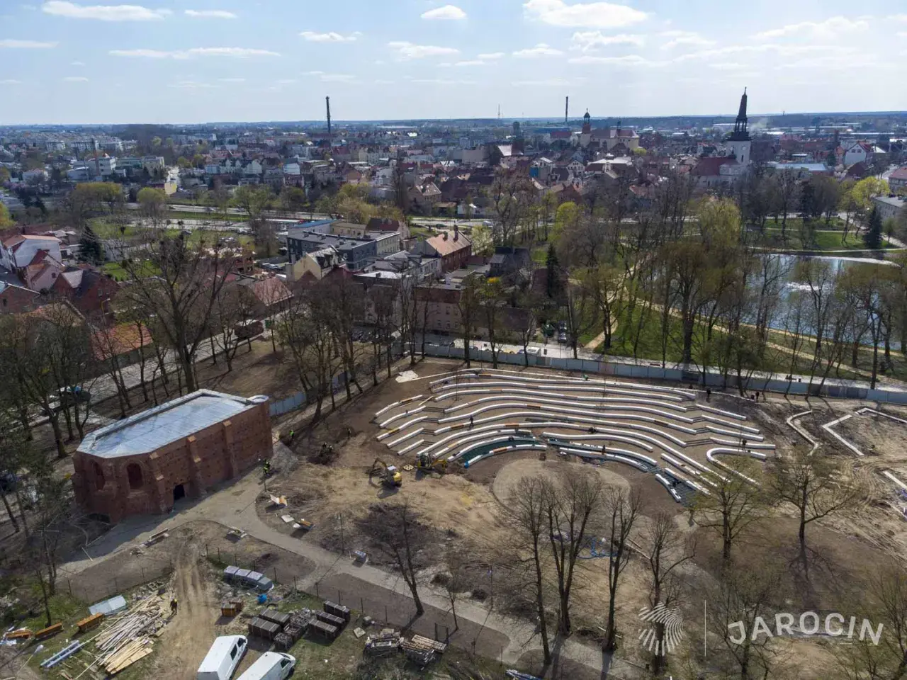 Nowe amfiteatralne trybuny w jarocińskim parku nabierają kształtów. W tle widać panoramę miasta i staw.