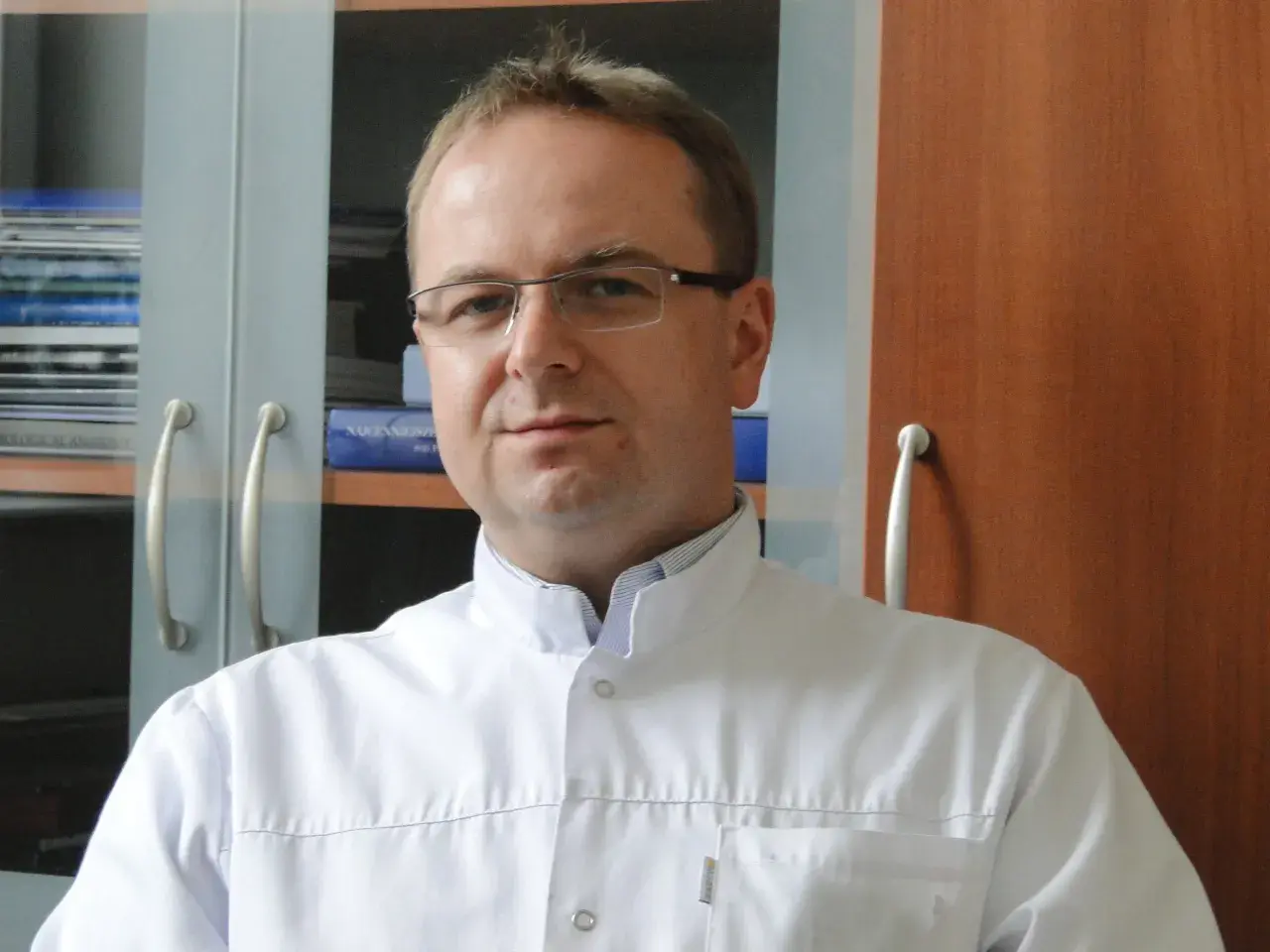 Portret mężczyzny w okularach i białym fartuchu lekarskim, na tle regałów z książkami.