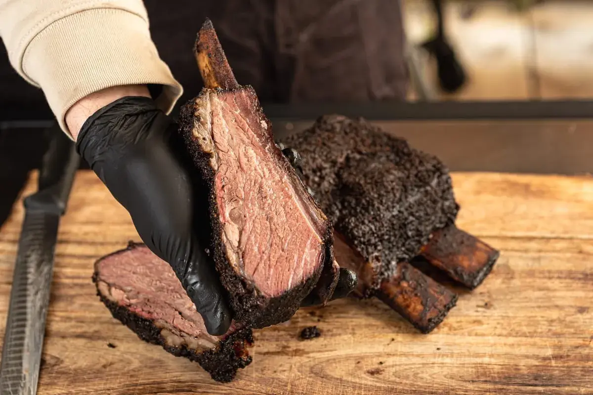 Une main gantée présente une côte de bœuf fumée, tendre et juteuse, sur une planche en bois.