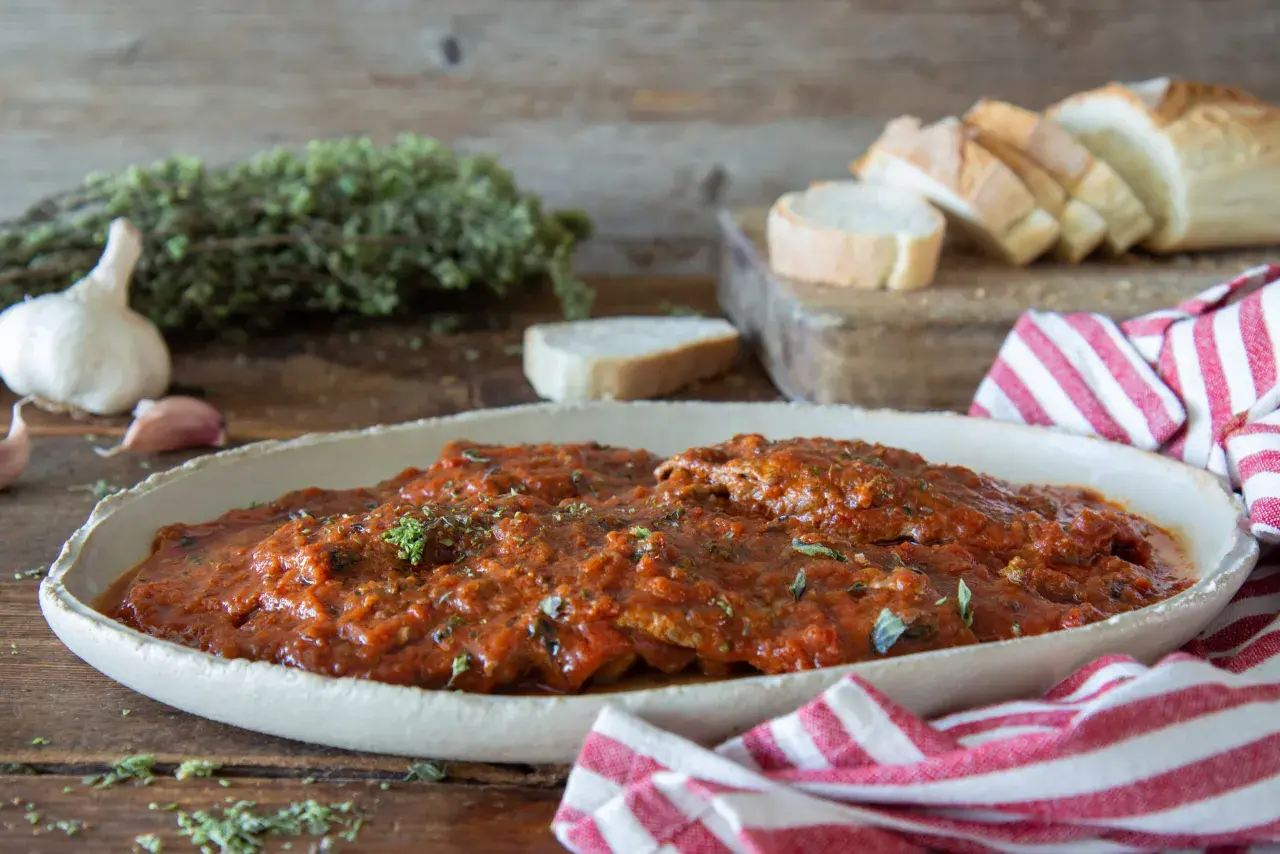 Herzhaftes Carne alla Pizzaiola in Tomatensauce, garniert mit Kräutern, neben Knoblauch, Brot und einem rot-weiß gestreiften Tuch.