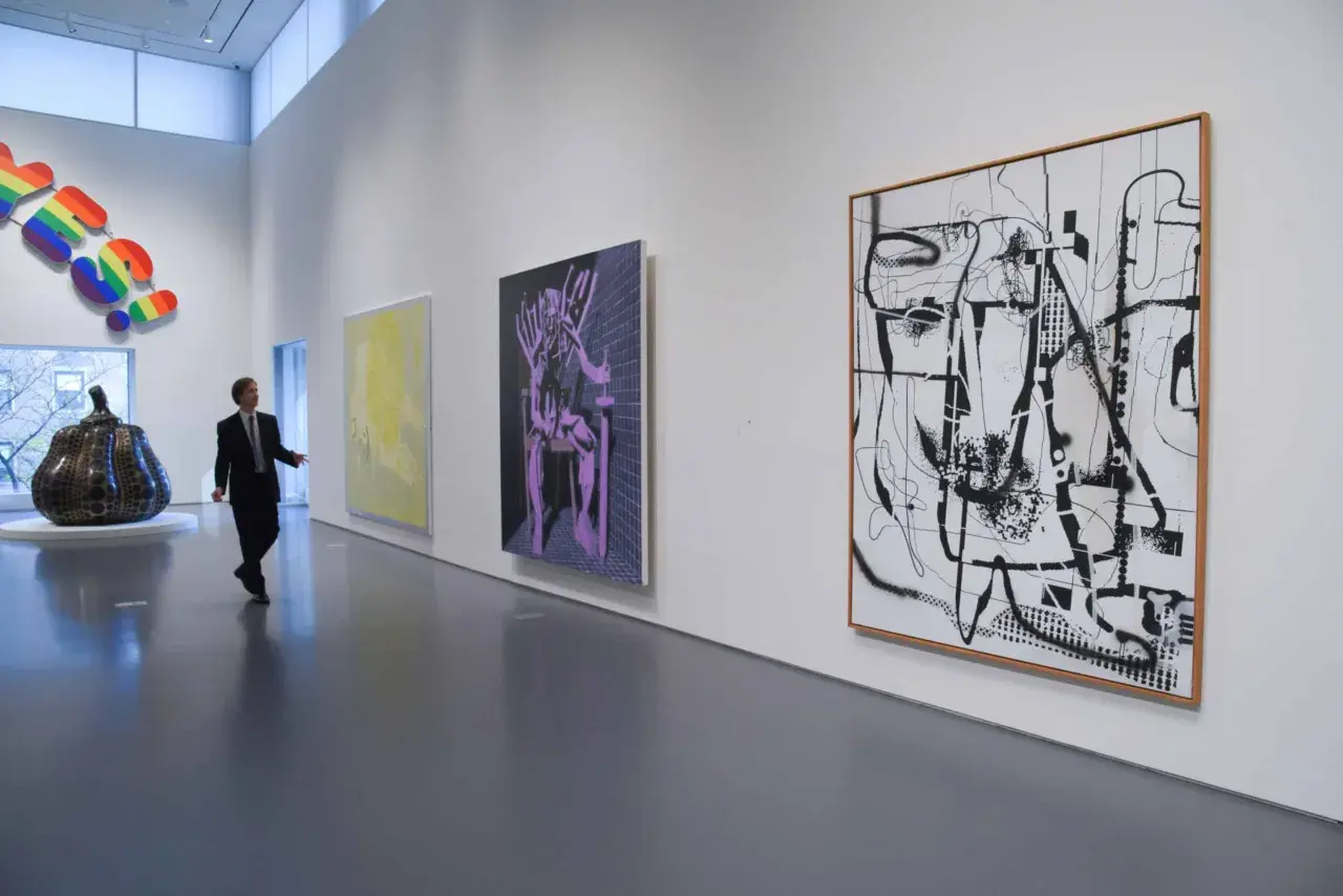 Un homme en costume observe une **analyse d'une œuvre d'art contemporain** dans une galerie. Des peintures abstraites et une sculpture colorée ornent les murs blancs.
