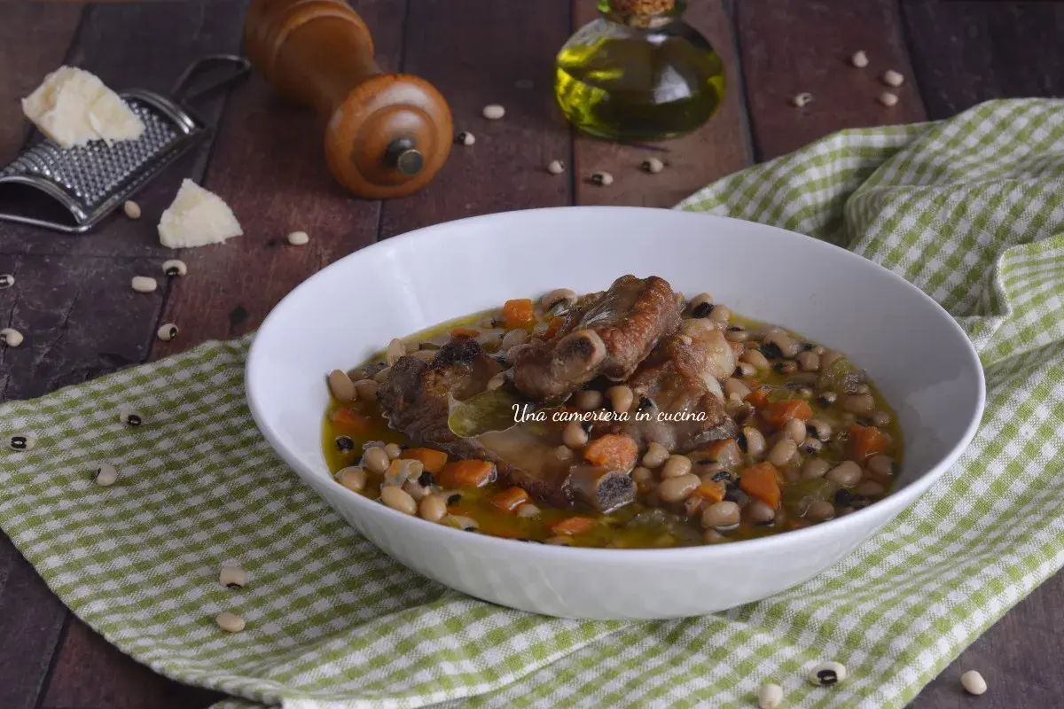Una ciotola di zuppa dei morti con fagioli, costine di maiale e carote, servita su una tovaglia a quadri verdi.