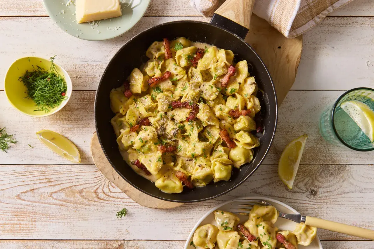 Pyszne tortellini grzybowe w kremowym sosie, posypane parmezanem i boczkiem, podane z cytryną i koperkiem.