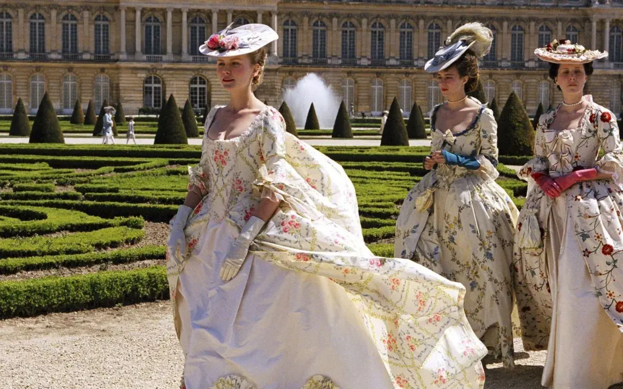 Kvinnor i magnifika **rococo fashion**-klänningar med blomstermönster promenerar i en formell trädgård framför ett palats.