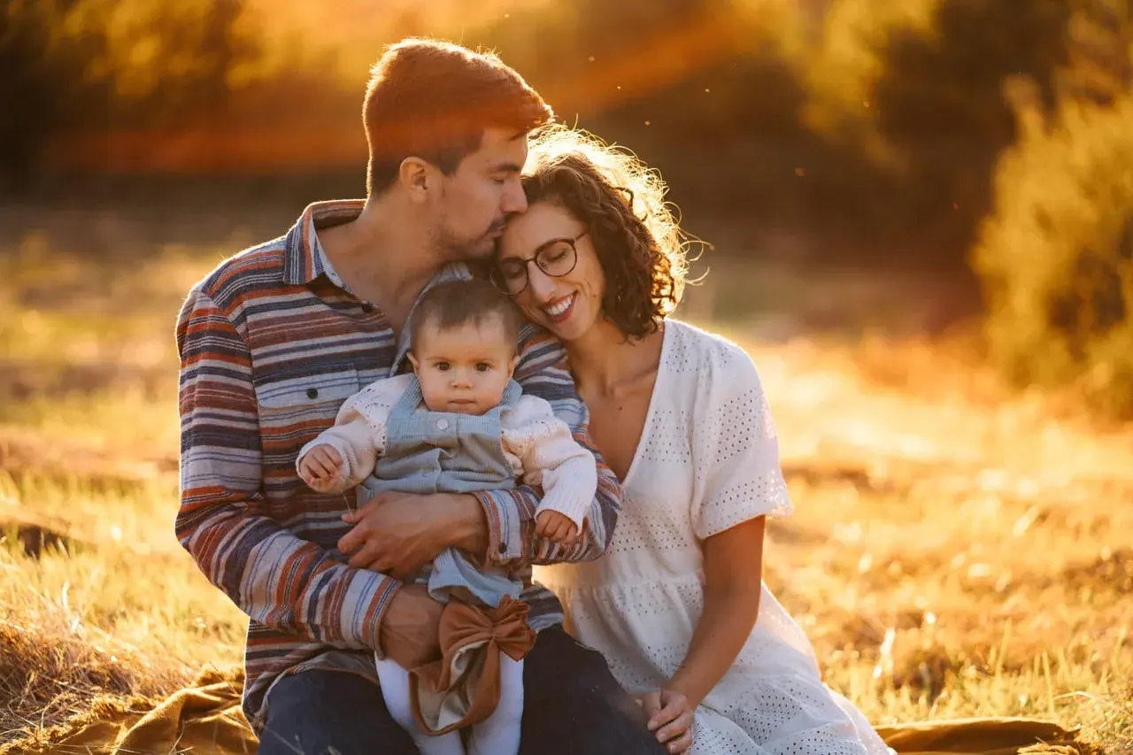 Ciepłe, złote światło otula rodzinę podczas sesji rodzinnych. Tata całuje główkę dziecka, mama uśmiecha się, wtulona w męża.