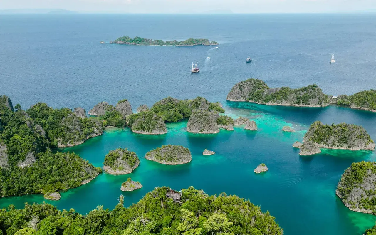 Traumhafte Anreise nach Raja Ampat: türkisfarbene Lagunen, grüne Inseln und Segelschiffe im azurblauen Meer.