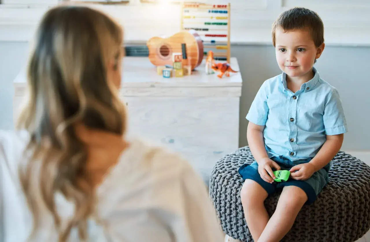 Chłopiec w niebieskiej koszuli siedzi na pufie, rozmawiając z psychologiem. W tle zabawki.