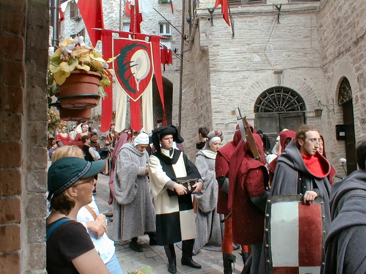 Persone in costume medievale sfilano in una strada acciottolata, con stendardi rossi e bianchi che sventolano. La scena evoca il **calendimaggio significato**, una festa di primavera.