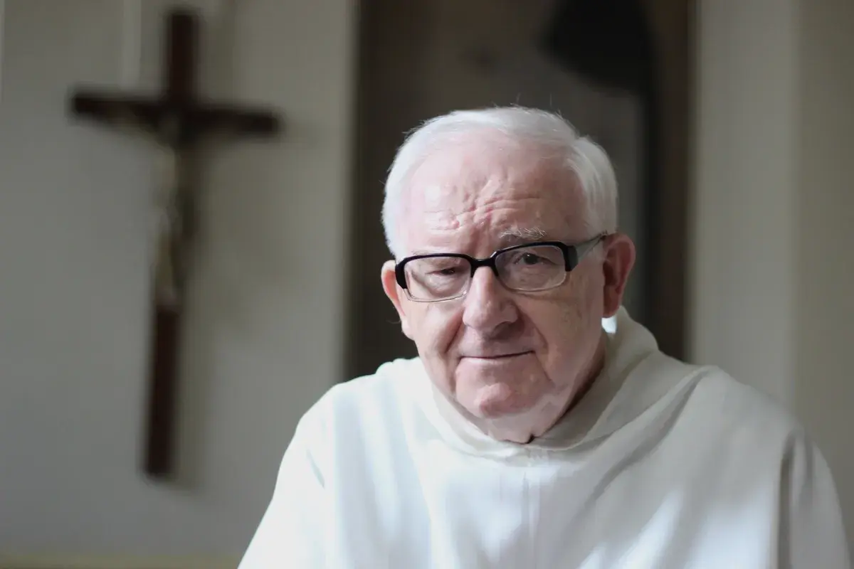 Starszy mężczyzna w okularach, w białej szacie, na tle krzyża. Może rozważa dogmat o nieomylności papieża.