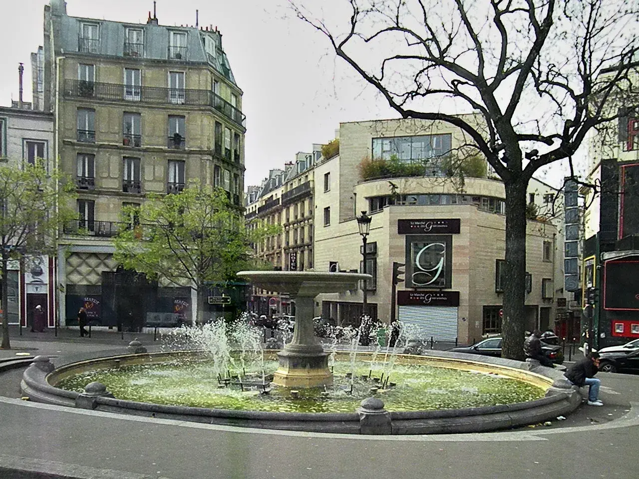 Fontanna na placu Pigalle w Paryżu, otoczona kamienicami i drzewami.
