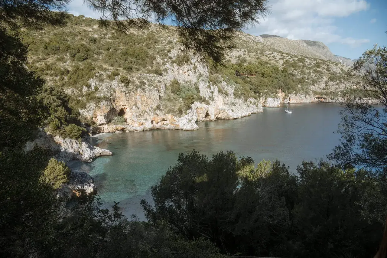 A serene view of Baia degli Infreschi, a secluded cove with clear turquoise waters, rocky cliffs, and lush greenery. A lone sailboat drifts peacefully in the bay.