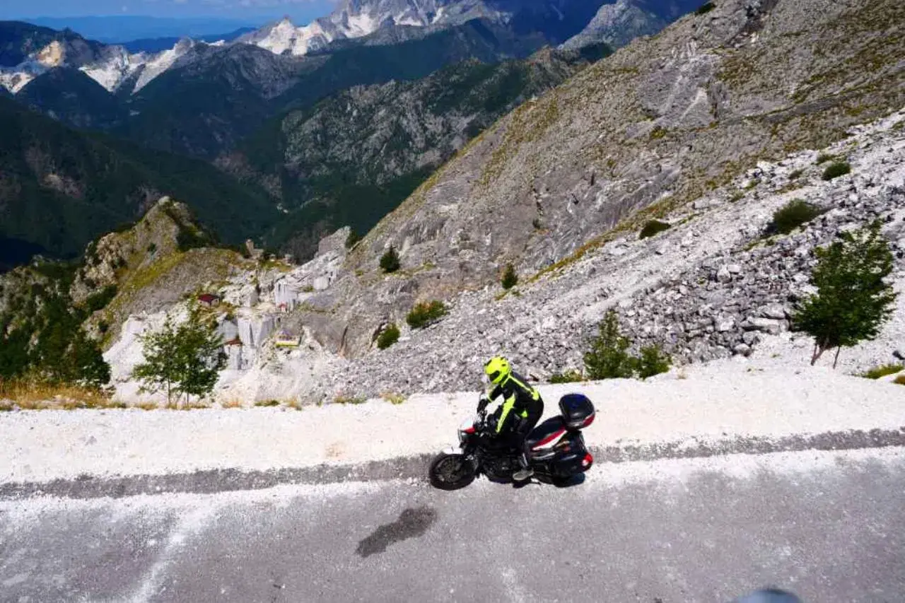 Un motociclista con casco giallo affronta il passo del vestito in moto, tra montagne rocciose e cave di marmo.