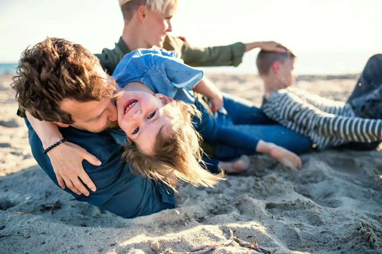 Rodzina na plaży, ojciec tuli córkę, która śmieje się do aparatu. W tle mama i syn. Idealne miejsce na jastrzębia góra atrakcje dla dzieci.