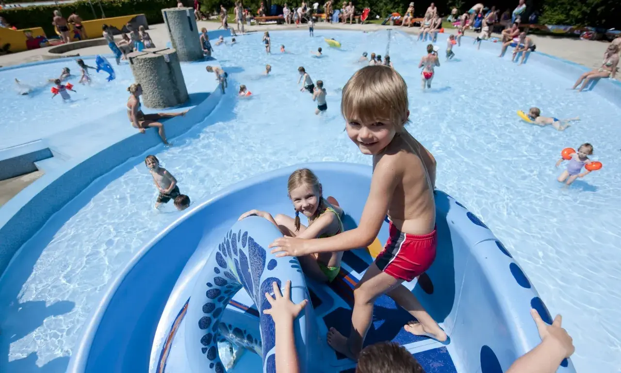 Fröhliche Kinder planschen und rutschen im Freibad. Ein Junge grinst in die Kamera, während ein Mädchen auf der blauen Rutsche sitzt. Ein perfekter Tag im bayerischen Wald mit Kindern.