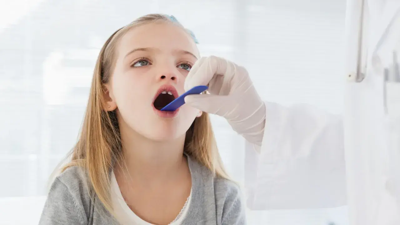 Niña con la boca abierta, examinada por un médico. Podría ser una consulta previa a una **operación amígdalas niños**.