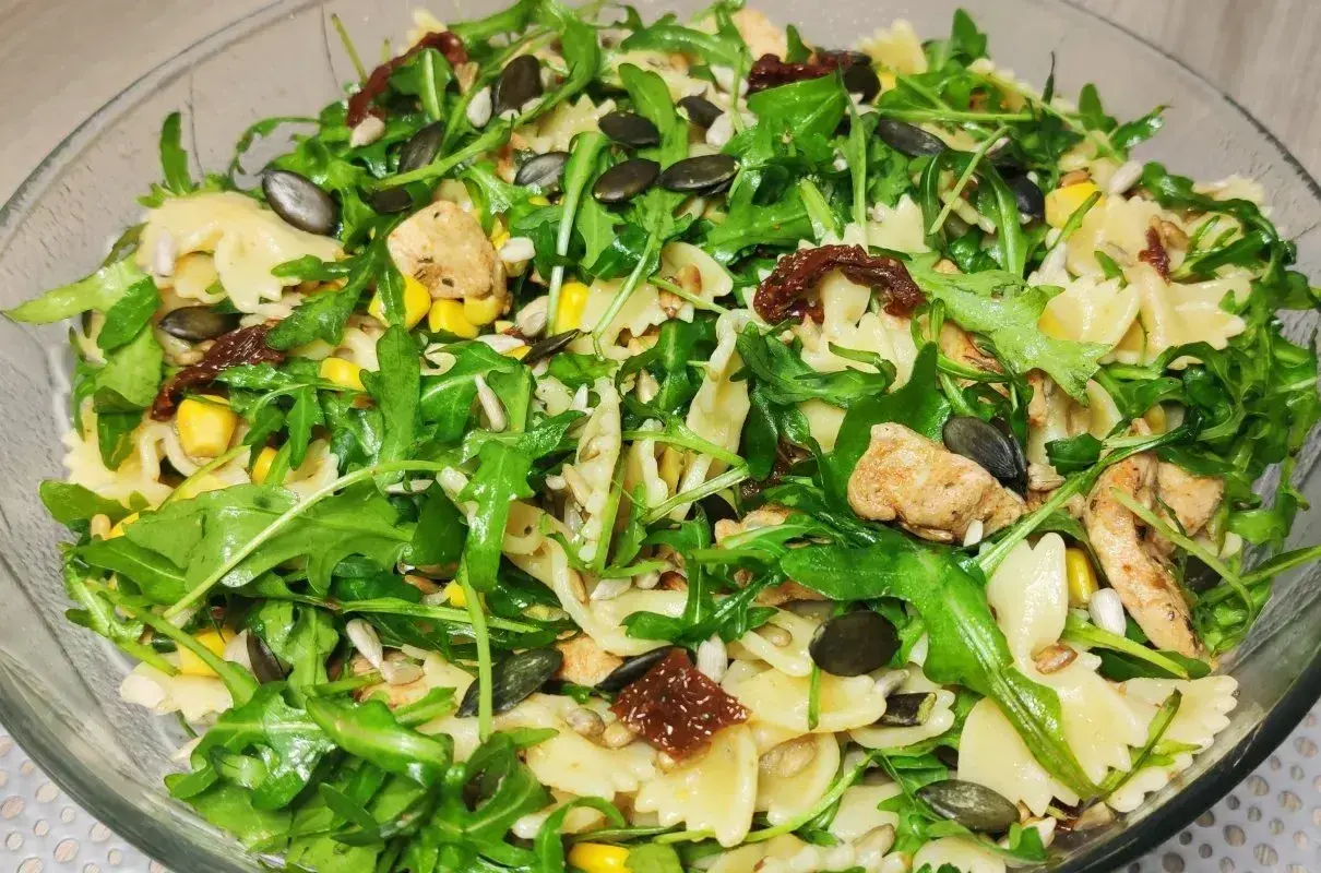 Pyszna sałatka z makaronem farfalle, rukolą, kurczakiem, kukurydzą i aromatycznymi suszonymi pomidorami. Posypana pestkami dyni i słonecznika.