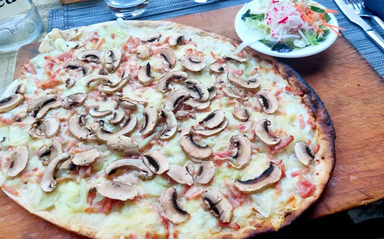 Ein köstlicher Straßburger Flammkuchen mit Pilzen und Speck, serviert auf einem Holzbrett, daneben ein kleiner Salat.